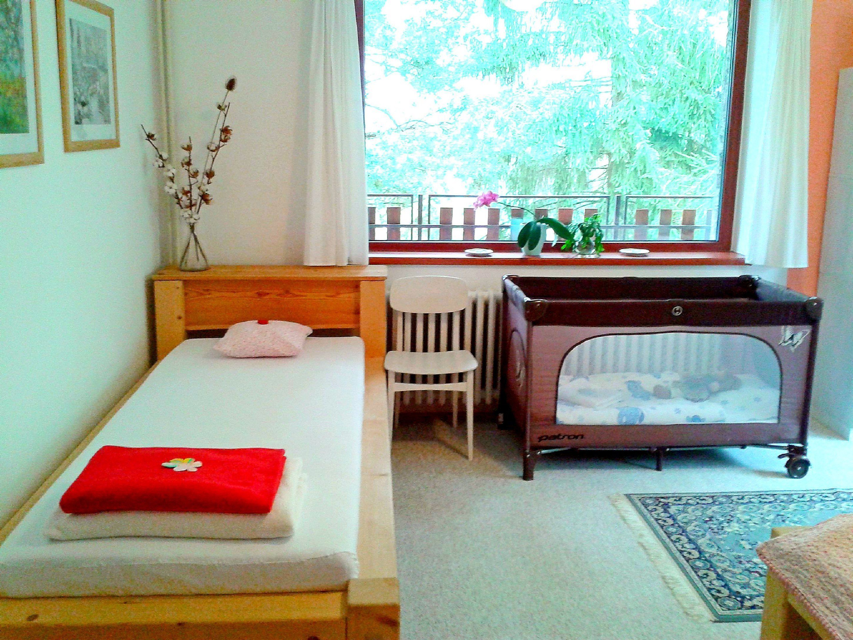 Un dormitorio con una cama individual, una mesita, una silla, una cuna y una ventana con vistas a zonas verdes.