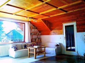 Une pièce chaleureuse avec un plafond en pente, une table en bois, un canapé avec des coussins, une bibliothèque et une fenêtre avec des rideaux.