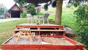 Une terrasse en bois avec une table de pique-nique et des chaises, entourée d’un jardin agrémenté d’un parterre surélevé et d’un arbre.