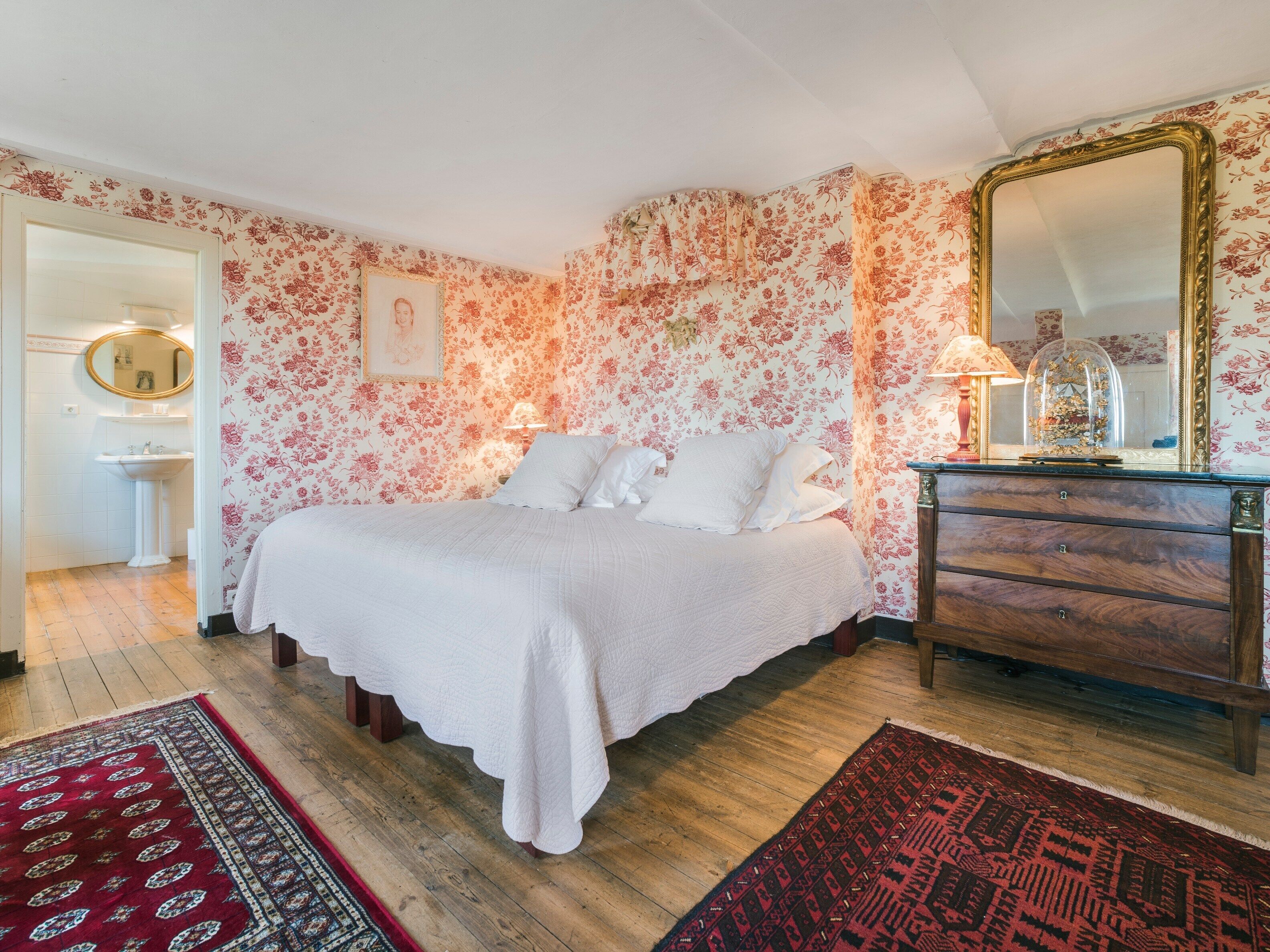 A bedroom with floral wallpaper, a bed with white linens, a wooden dresser, and a mirror.