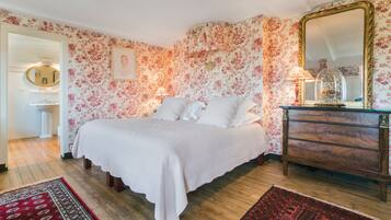 A bedroom with floral wallpaper, a bed with white linens, a wooden dresser, and a mirror.