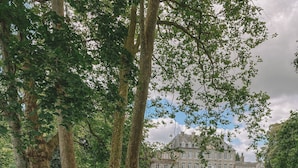 A person in a green dress with a straw hat and a woven bag standing in a garden with hydrangeas and a large building in the background. - Château de Boucéel (Saint-James)