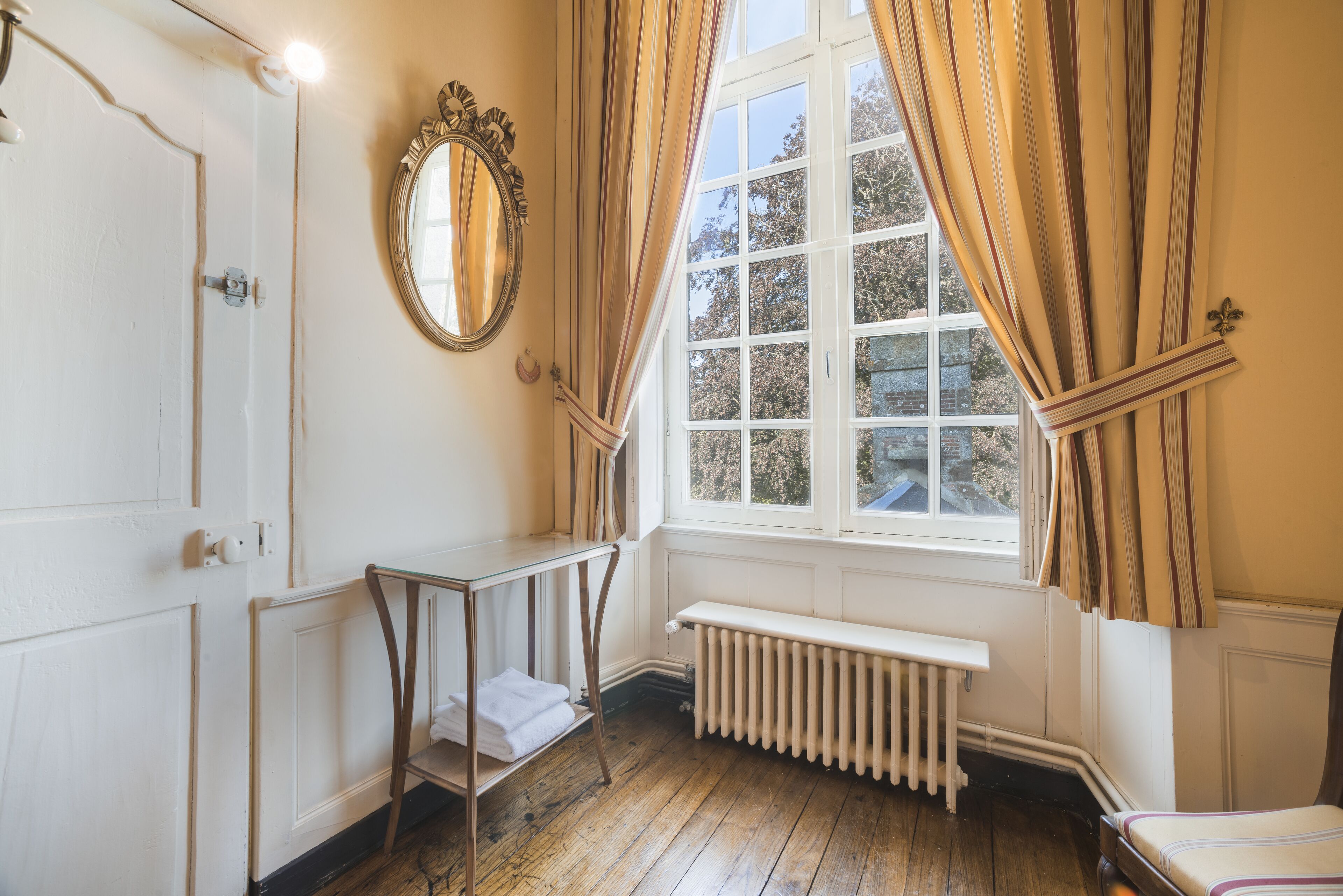 Ein Zimmer mit Holzboden, einem weißen Heizkörper, einer Bank, einem Spiegel und einem Fenster mit Vorhängen.