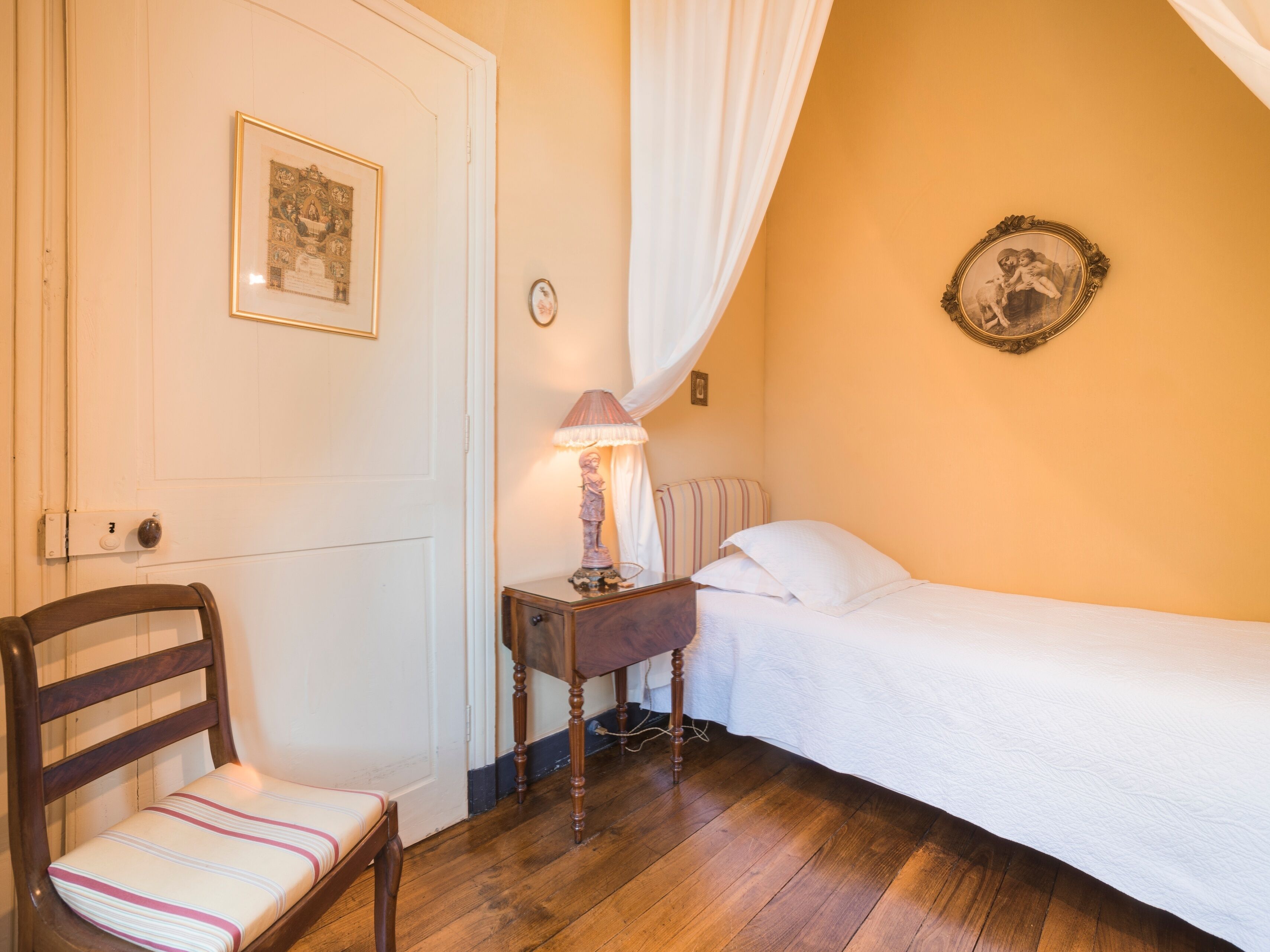 A bedroom with a wooden floor, a bed with white bedding, a chair with striped cushions, a bedside table with a lamp, and a framed picture on the wall.