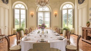A formal dining room with a long table set for a meal, surrounded by chairs, and large windows offering a view of the outdoors. - Château de Boucéel (Saint-James)