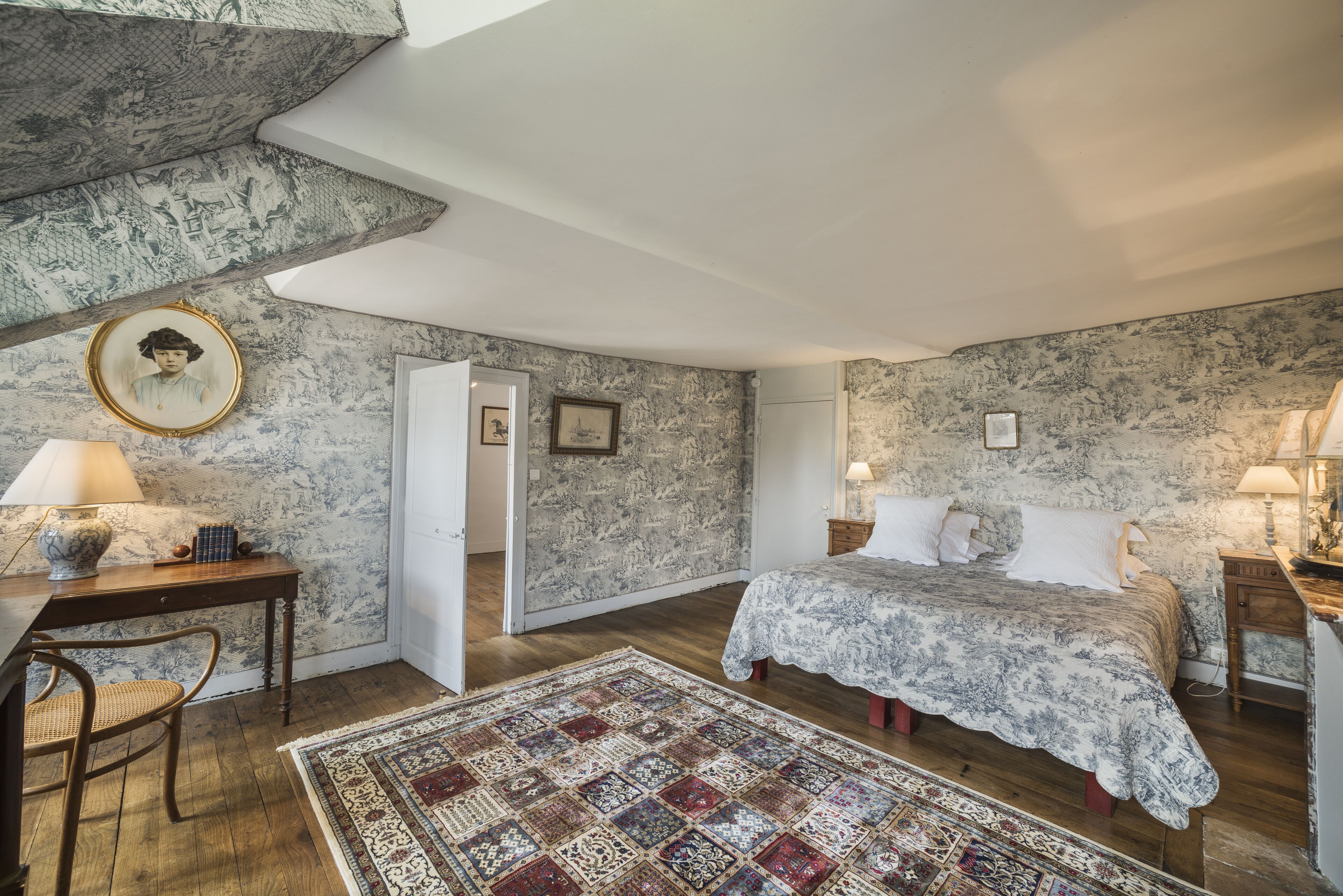 Ein Schlafzimmer mit einem Bett, einem Schreibtisch, einem Stuhl, einem Teppich und einer Tür zu einem anderen Raum.