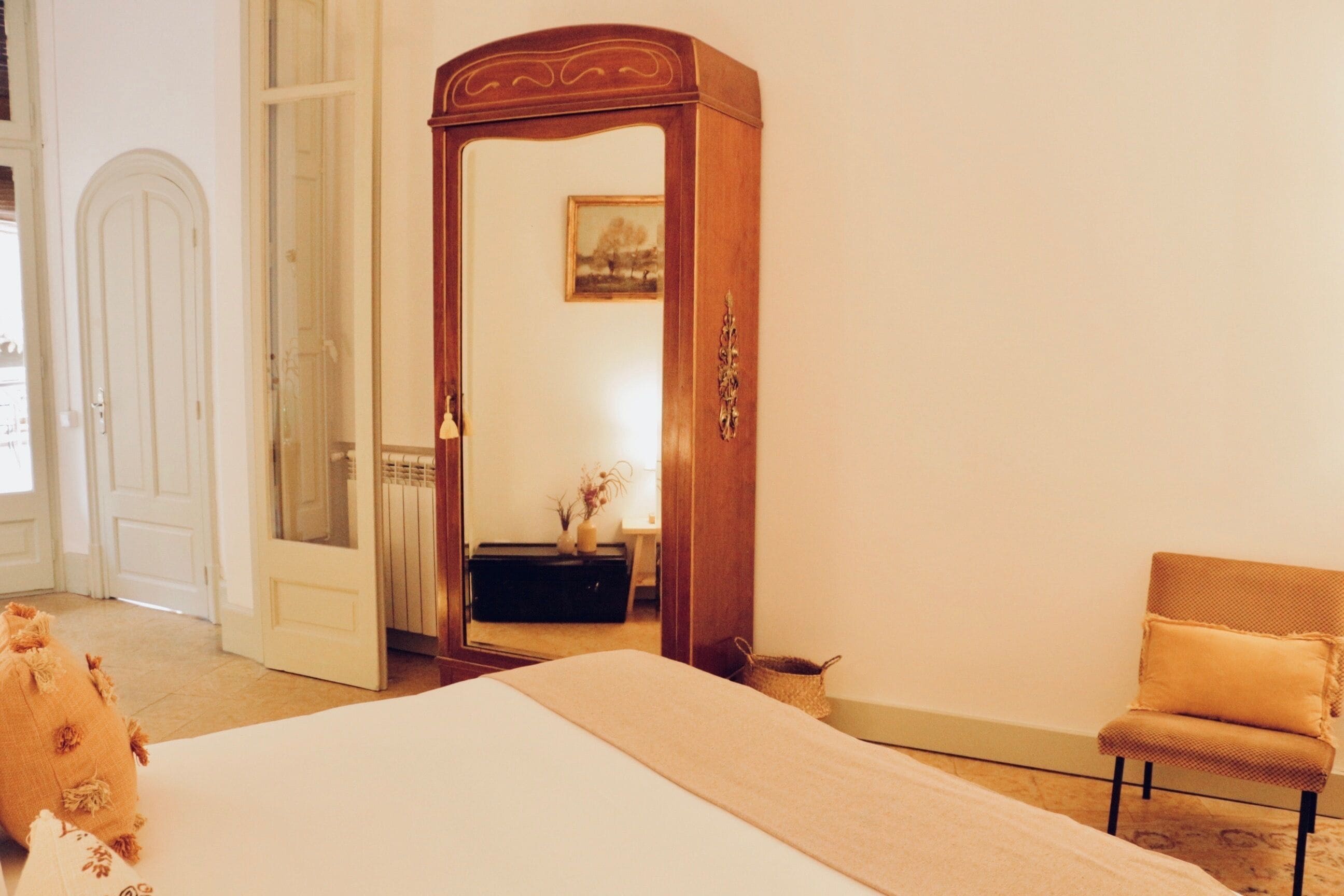 A bedroom with a large bed, a wooden wardrobe, a chair, and a door leading to another room.