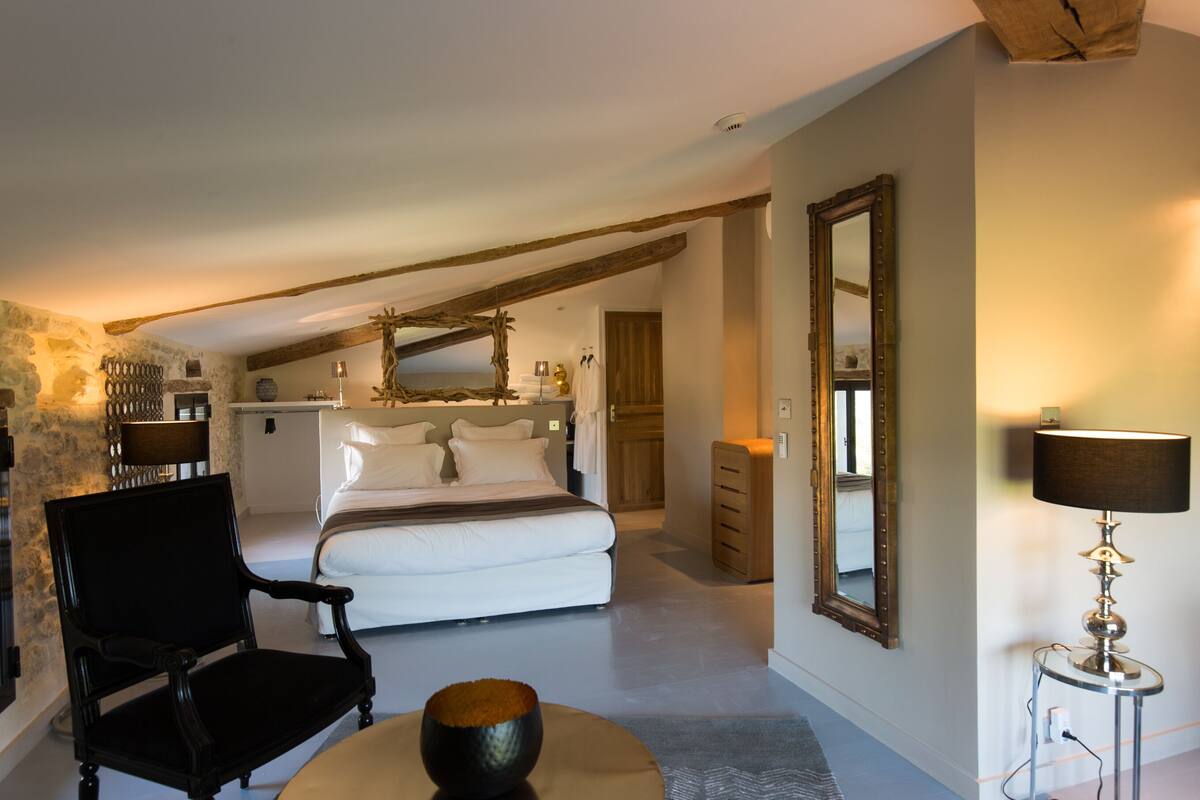 Ein Schlafzimmer mit einem großen Bett, einem Holzstuhl, einem runden Tisch mit einer Schale, einem Spiegel und einer Lampe.