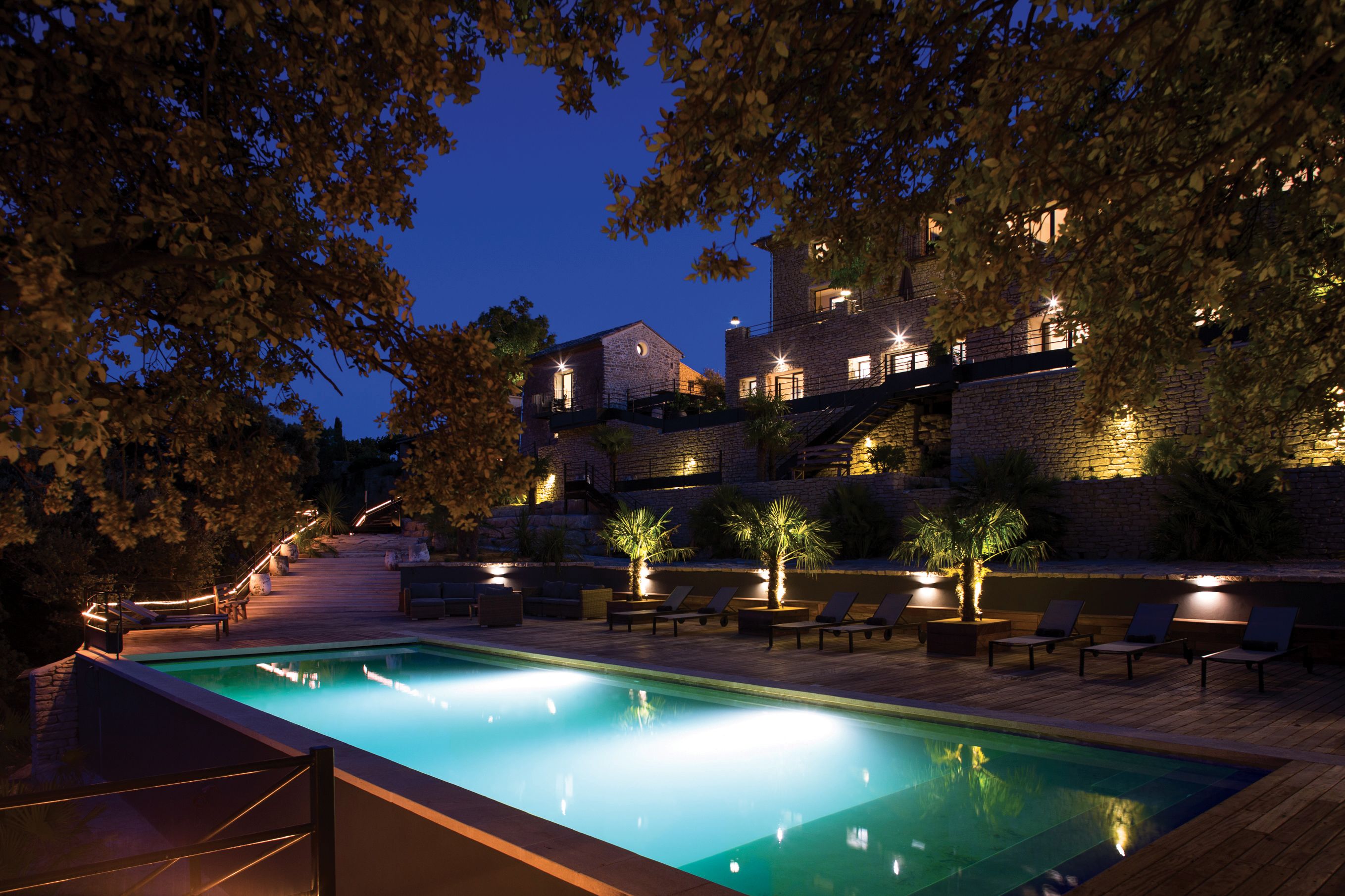 A swimming pool with lounge chairs and a wooden deck, surrounded by trees and a stone building with illuminated windows.