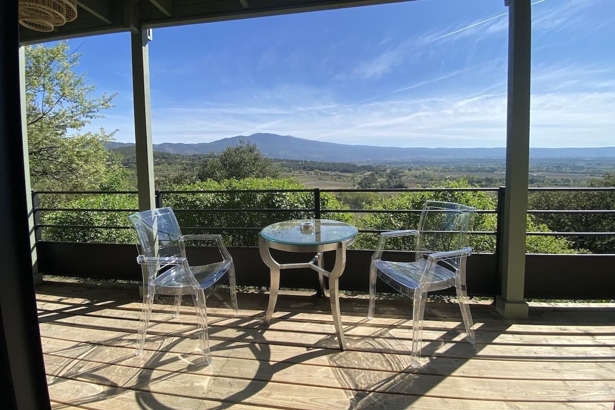 Ein überdachter Außenbereich mit Tisch und Stühlen, der einen malerischen Ausblick auf eine Landschaft mit Bergen und Bäumen bietet.