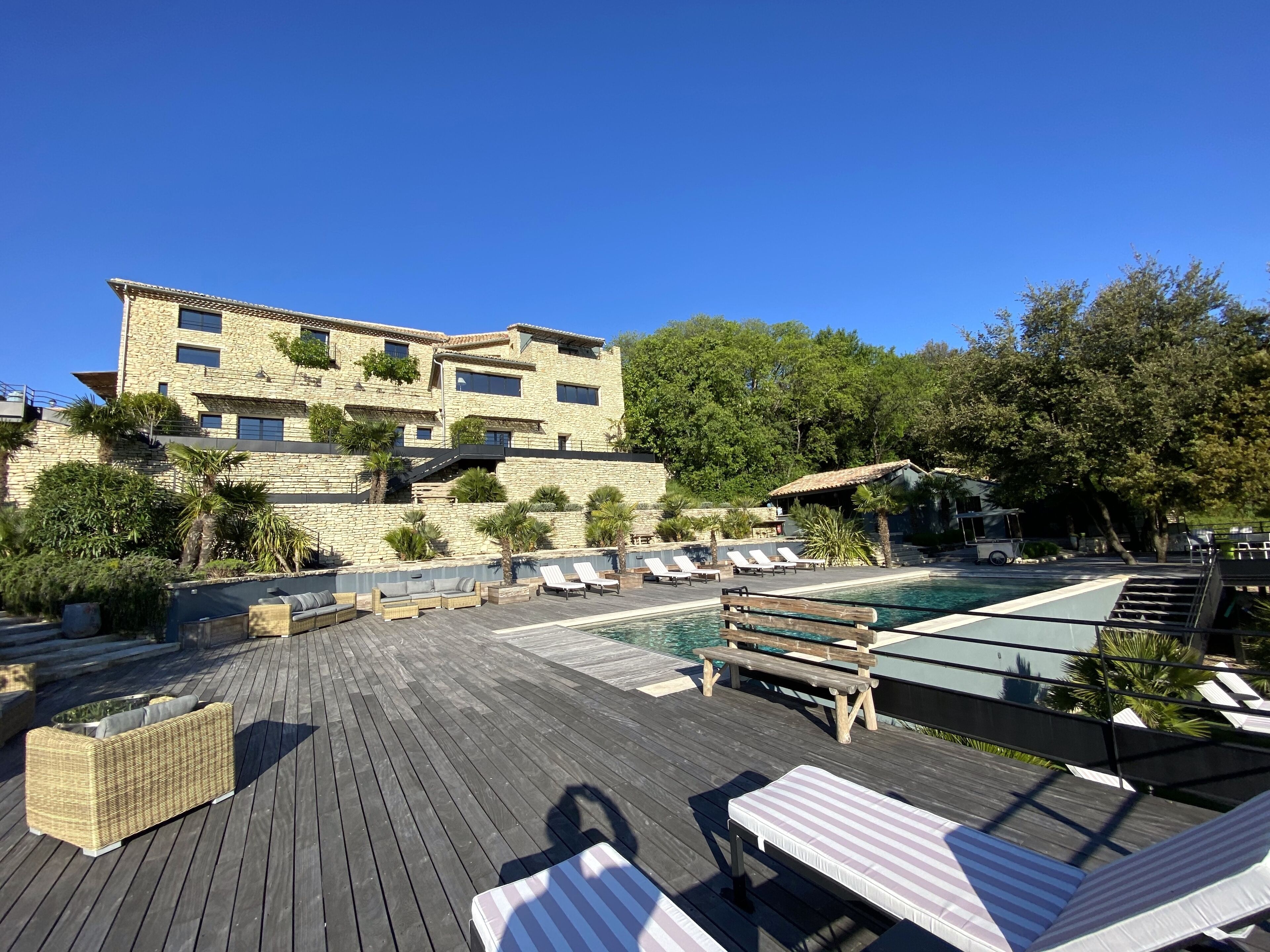 Une terrasse en bois avec des chaises longues et une piscine, entourée de bâtiments en pierre et de verdure.