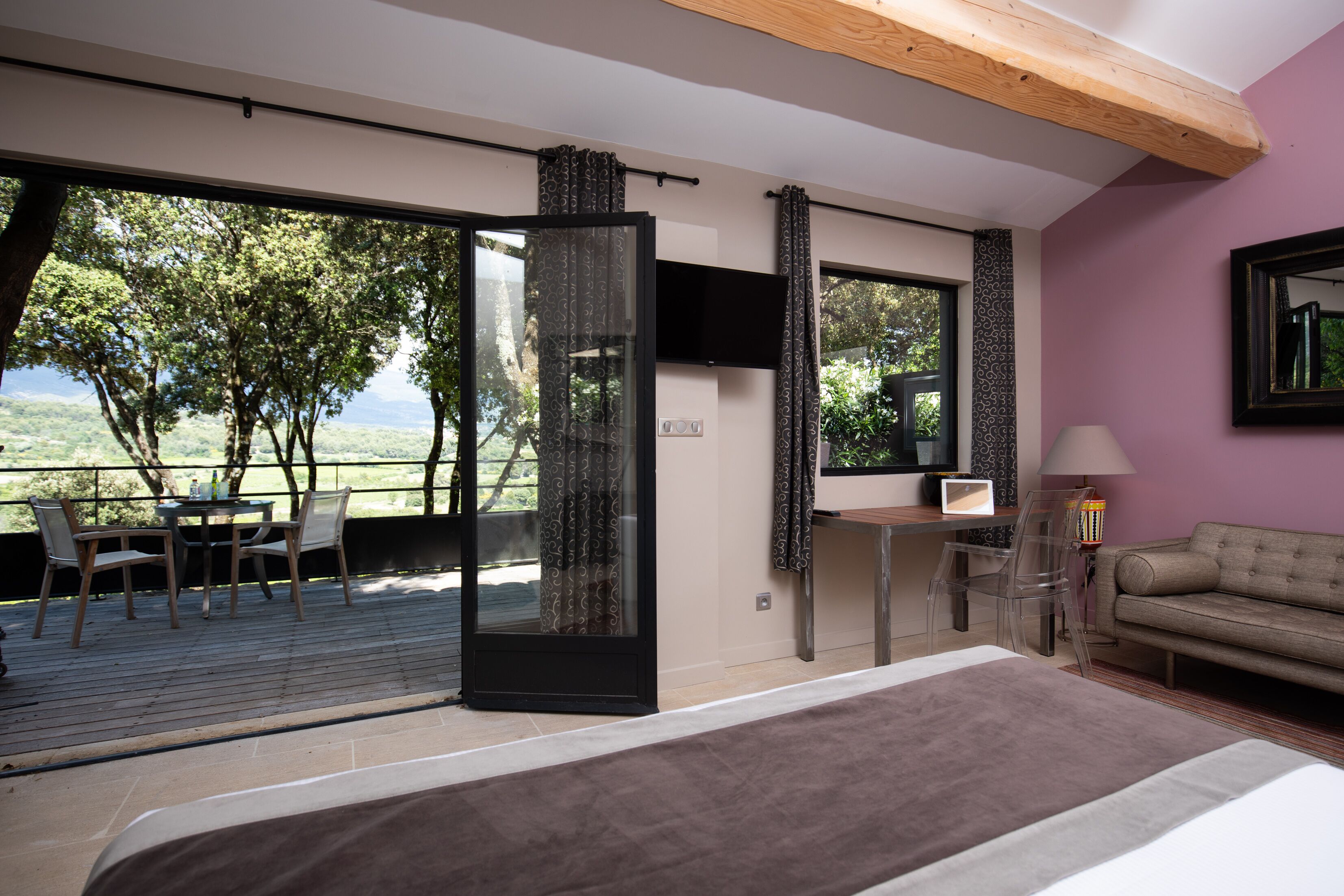 Une chambre avec un grand lit, une télévision, un canapé et un bureau avec une chaise. Il y a un balcon avec une table et des chaises, et une vue sur les arbres et les montagnes.