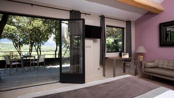 Une chambre avec un grand lit, une télévision, un canapé et un bureau avec une chaise. Il y a un balcon avec une table et des chaises, et une vue sur les arbres et les montagnes.