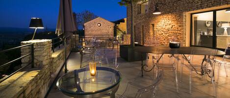 An outdoor terrace with stone walls, clear glass tables, and chairs, featuring a stone building in the background.