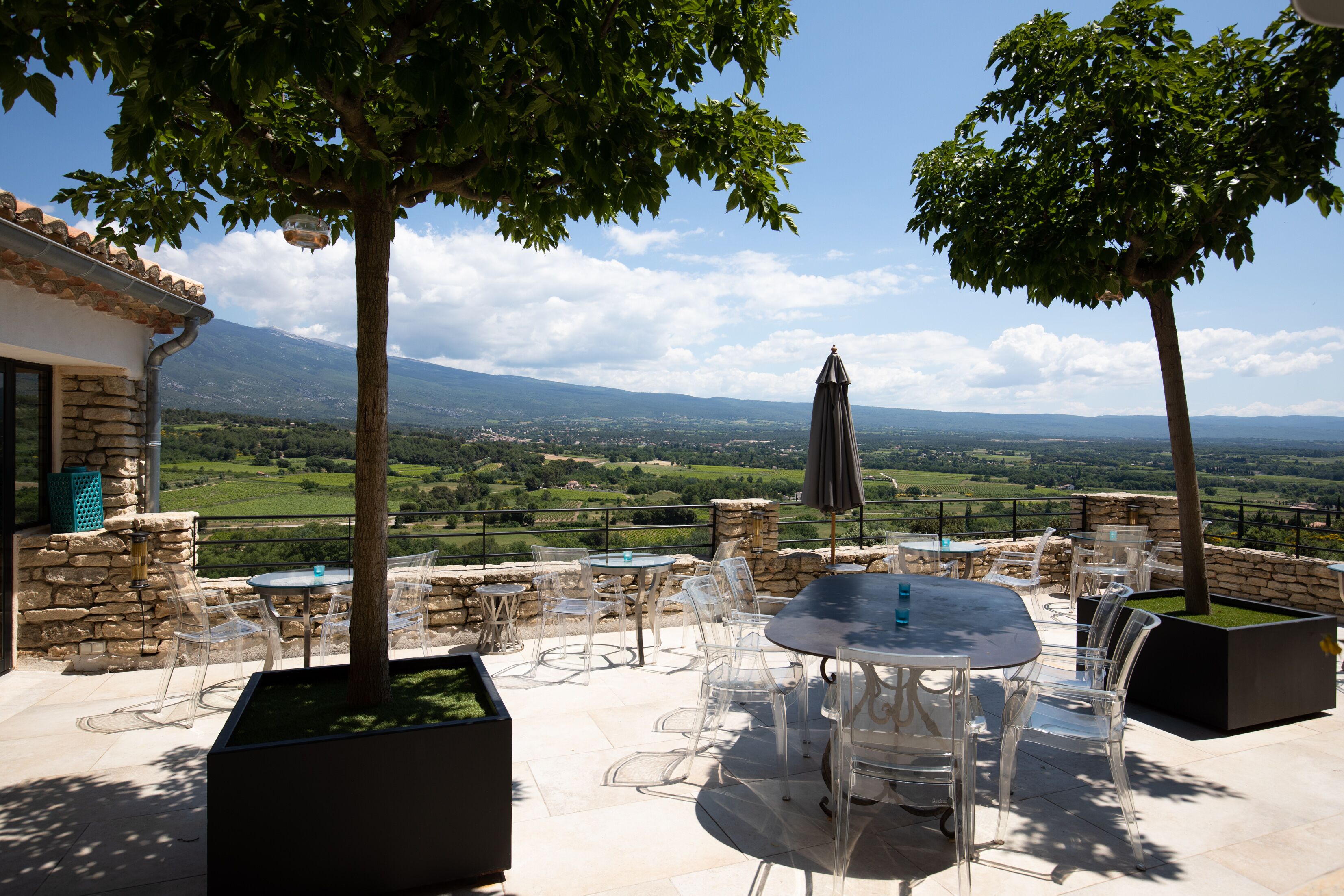 An outdoor patio with clear chairs and tables, a stone wall, and a view of a green landscape and mountains in the distance.