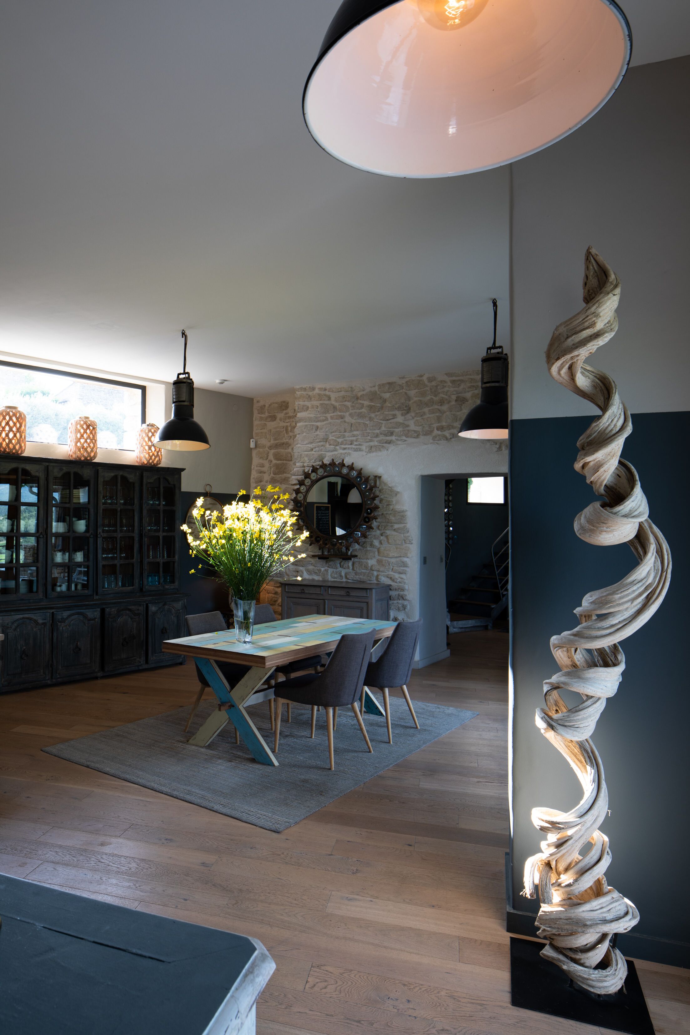 A modern kitchen with a dining area, featuring a spiral sculpture, a large window, and a stone wall.
