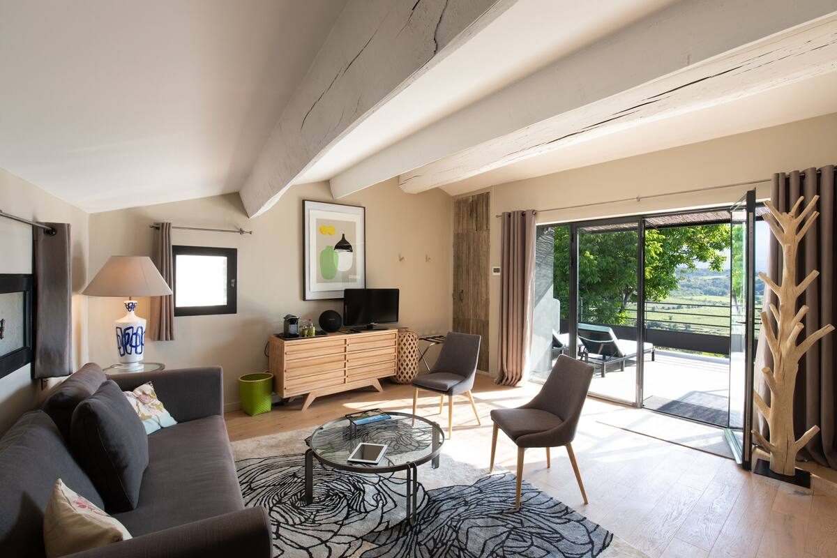 Ein modernes Wohnzimmer mit einer Couch, Sesseln, einem Couchtisch, einem Fernseher und einem großen Fenster mit Blick ins Freie.