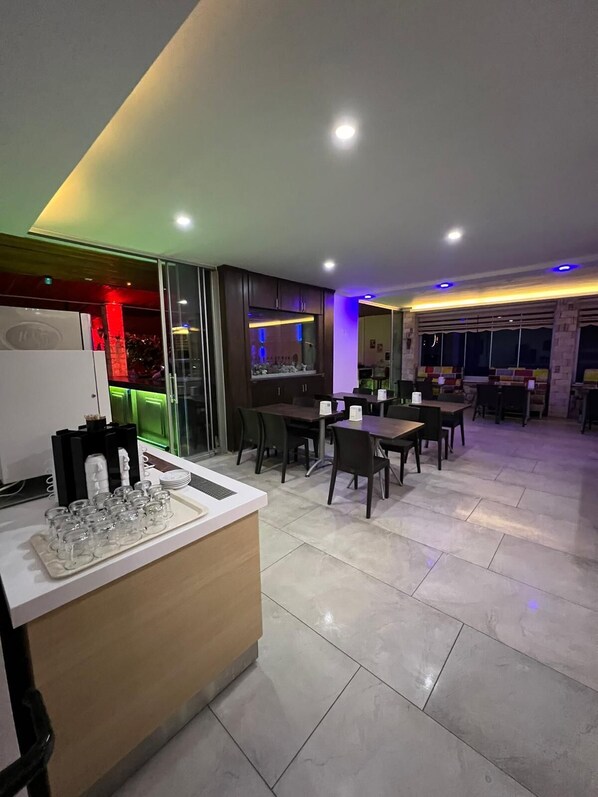 A modern dining area with a central counter, multiple tables, and a large window providing a view of the night sky. - Miray Hotel (Marmaris)