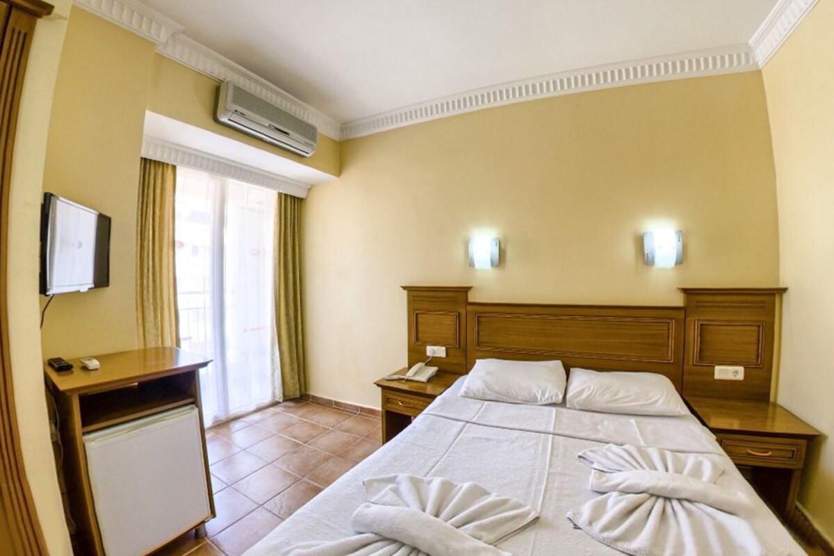 A hotel room with a bed, bedside tables, a desk, a chair, an air conditioner, a TV, and a window with curtains.