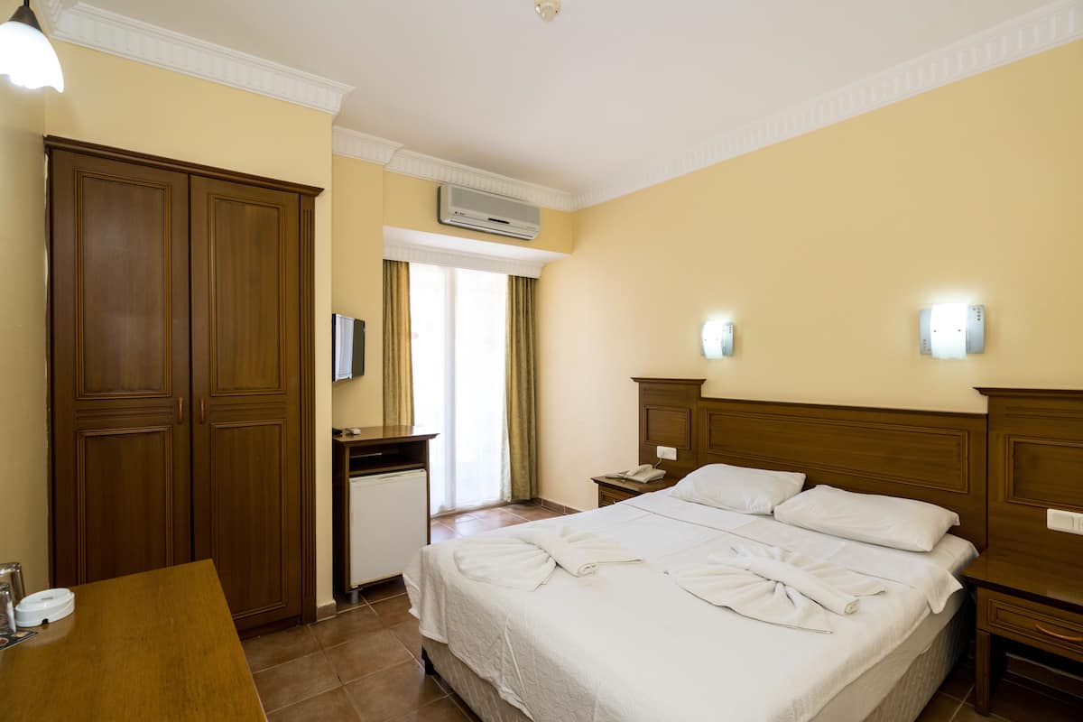 A hotel room with a large bed, a wooden wardrobe, a desk with a chair, a bedside table, and a wall-mounted air conditioning unit.