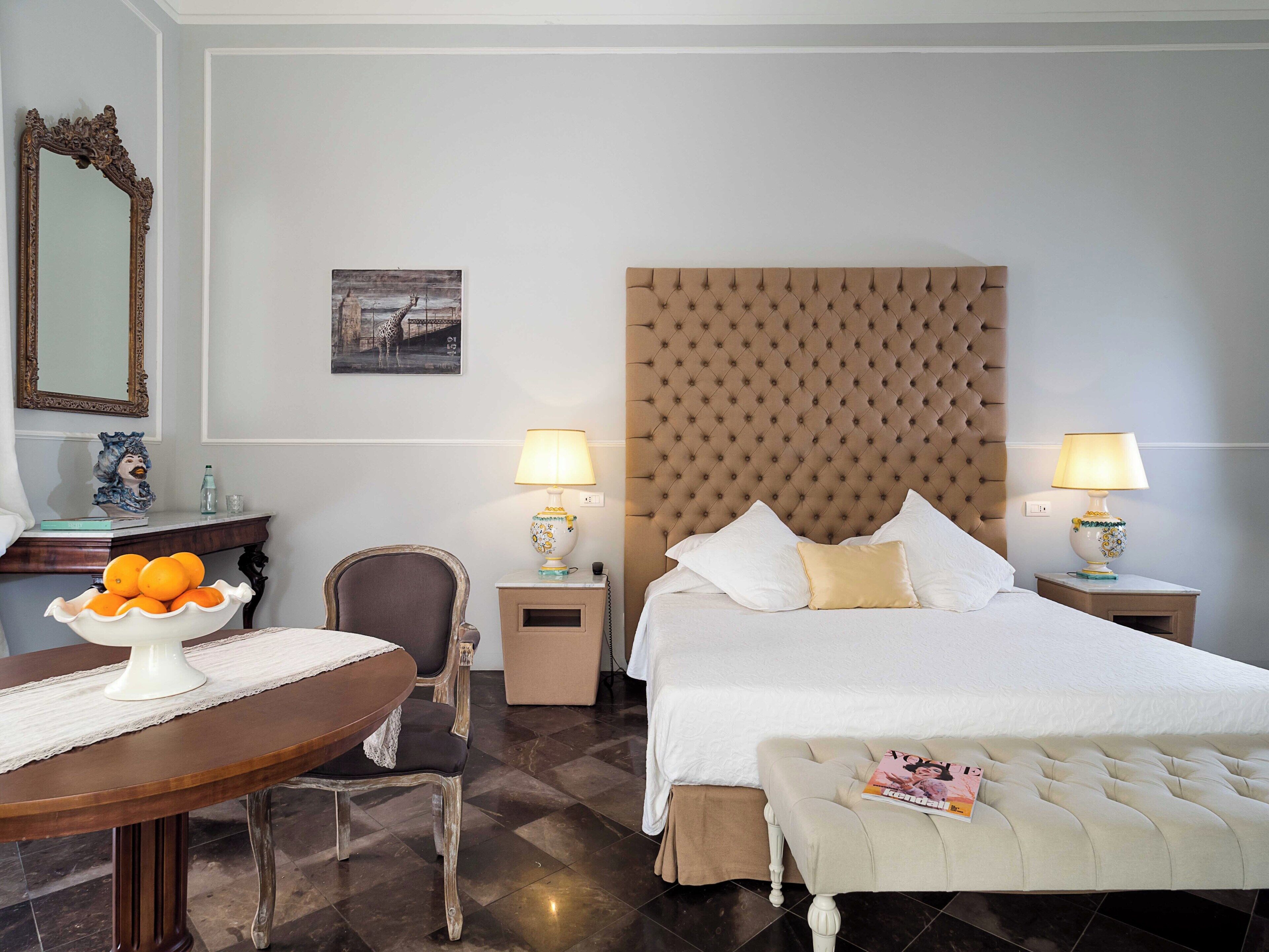 Habitación de hotel con una cama grande, dos lámparas de noche, una silla, una mesa con un frutero y un cuadro en la pared.