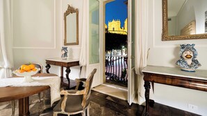 Una habitación con vista a un edificio amarillo, una silla de madera, una mesa pequeña con un cuenco de fruta y un busto decorativo.
