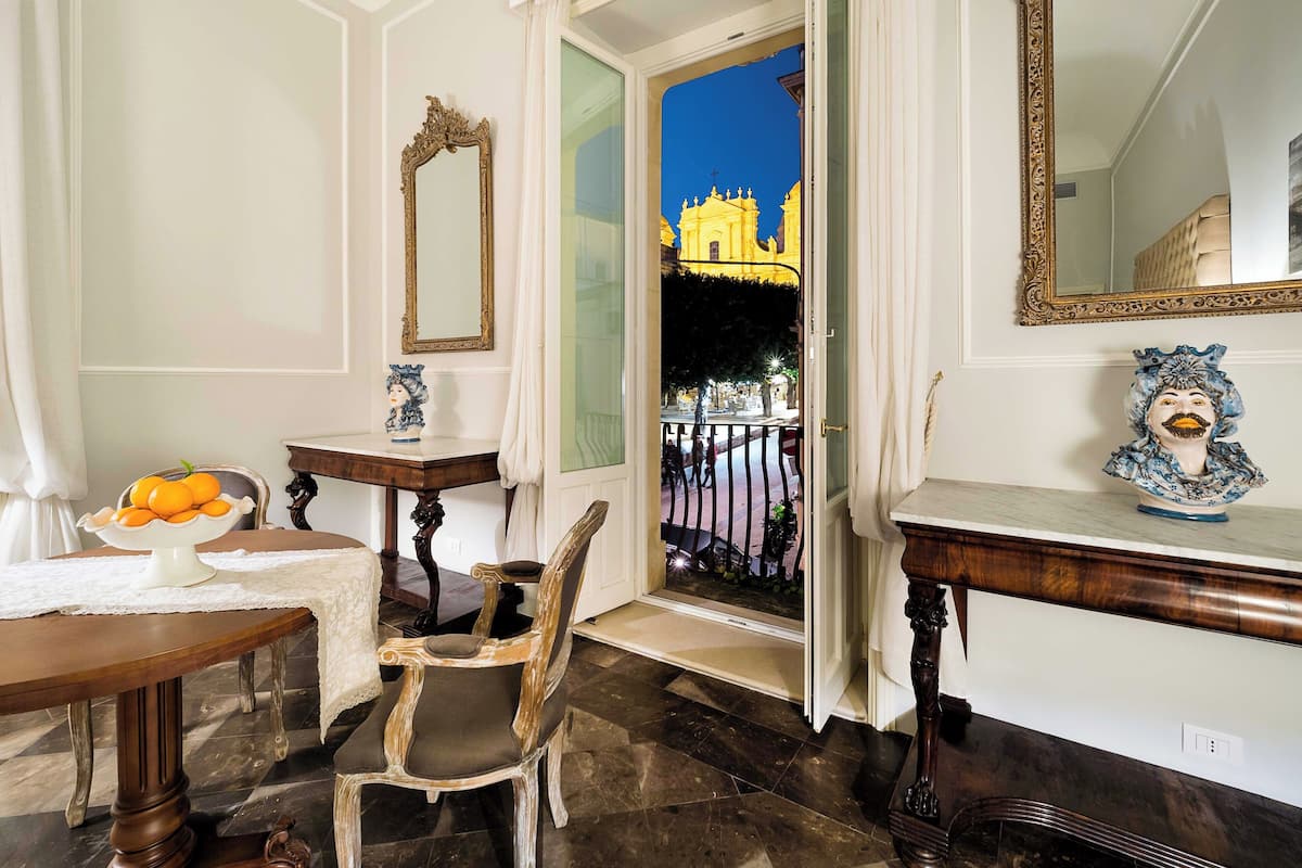 A room with a view of a yellow building, a wooden chair, a small table with a bowl of fruit, and a decorative bust.