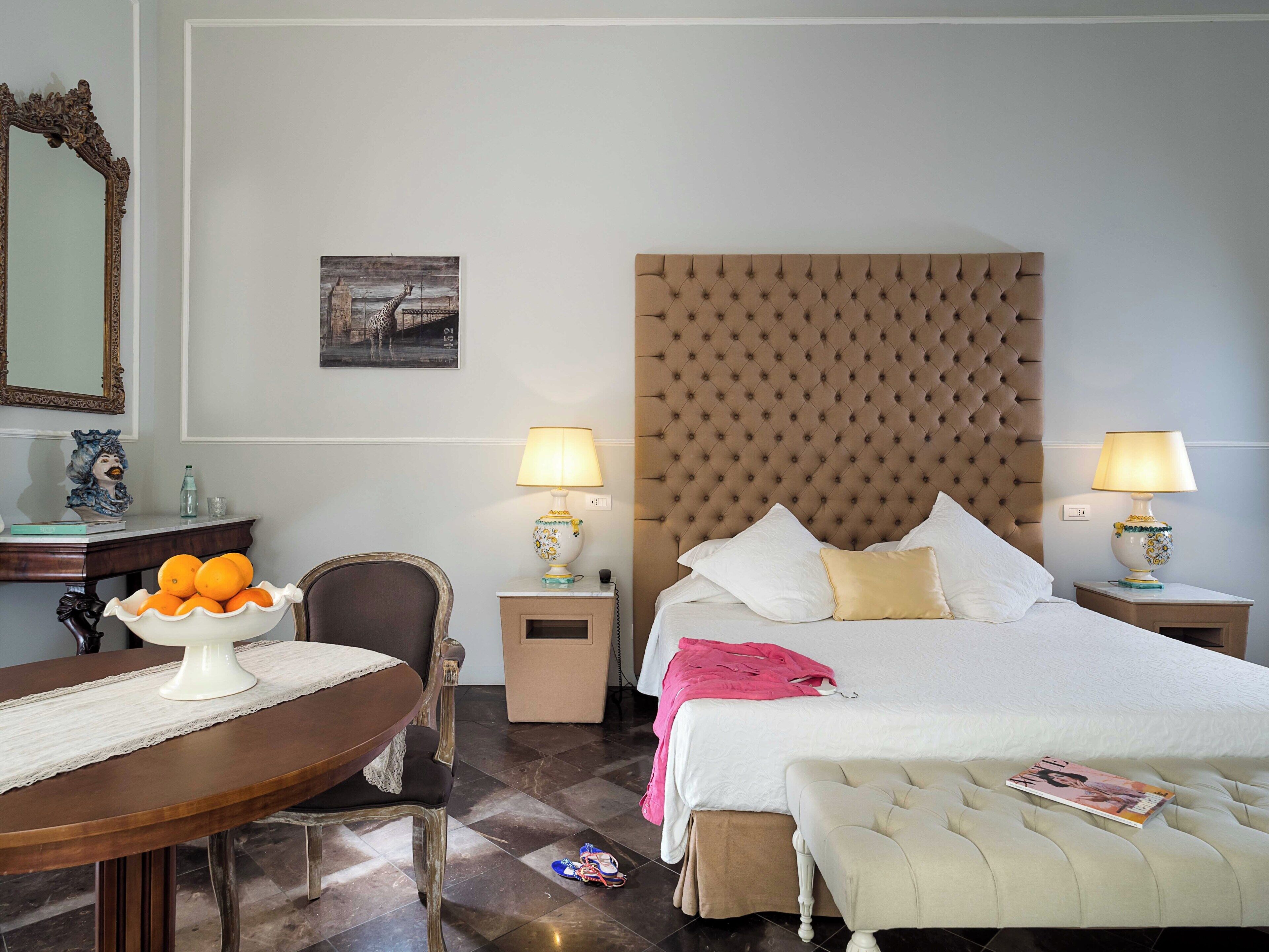 Habitación de hotel con una cama grande, dos mesitas de noche con lámparas, una silla, un escritorio con un frutero, un espejo y un cuadro en la pared.