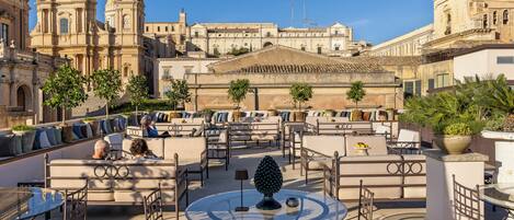 Una terraza en la azotea con mobiliario exterior, un árbol decorativo y vistas a edificios históricos.