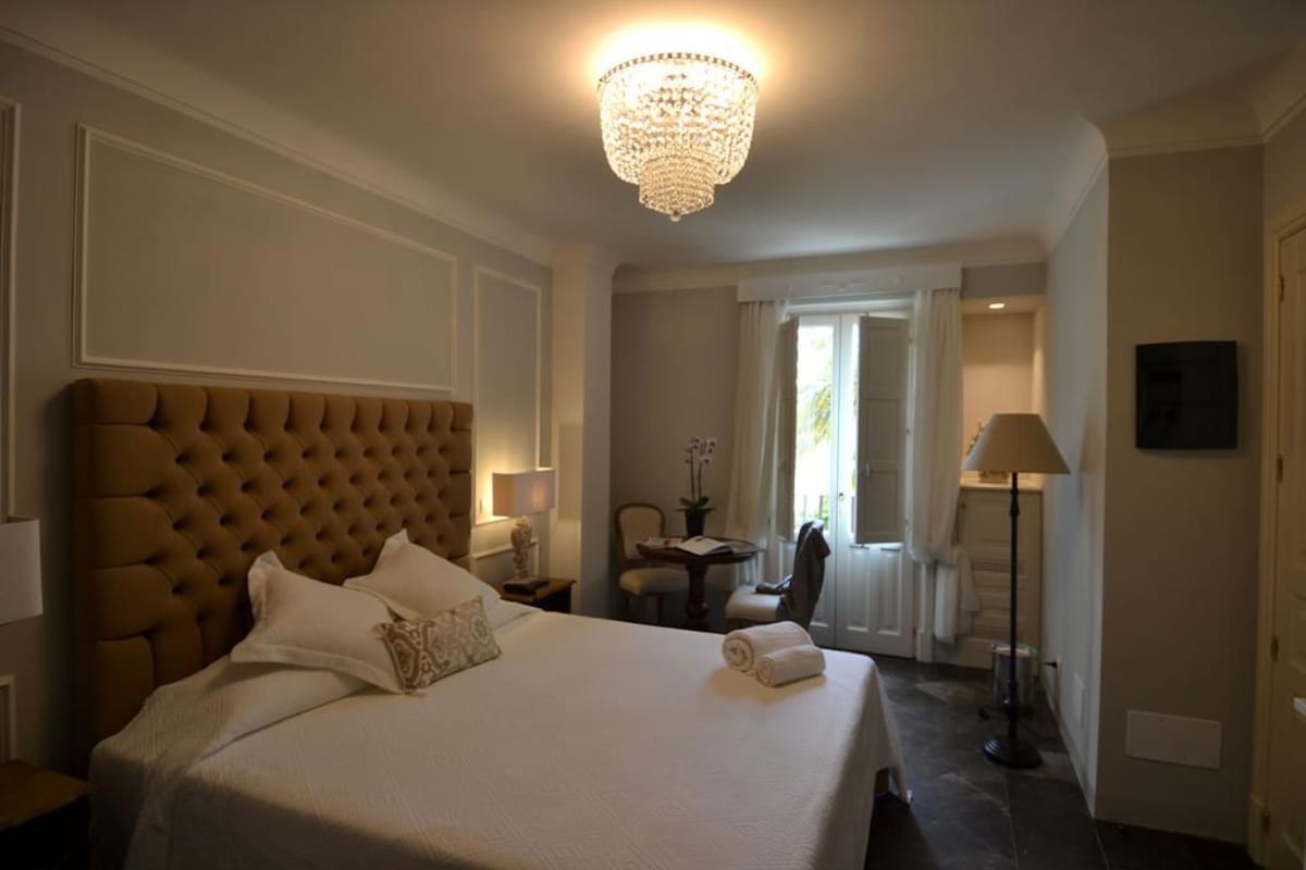 A hotel room with a large bed, a chandelier, a desk with a lamp, and a view of the outdoors.