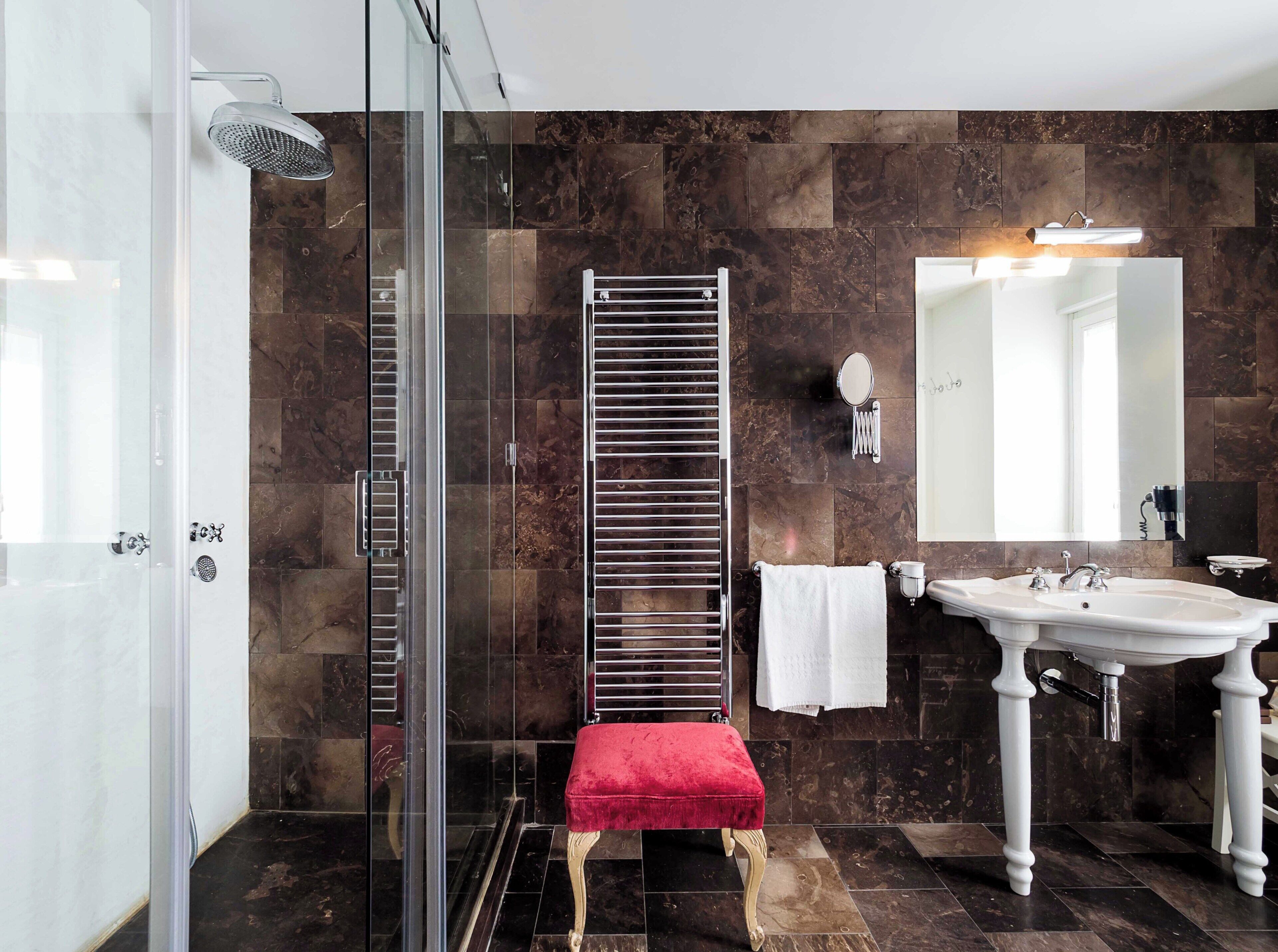 Een moderne badkamer met een glazen douchecabine, een hoge verticale verwarming, een rode bank, een witte wastafel en een grote spiegel.