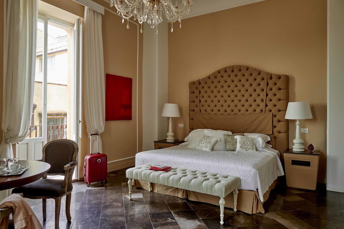 A hotel room with a large bed, two bedside tables with lamps, a bench, a chair, a dining table with a tray, and a view of the outside through a window with curtains.