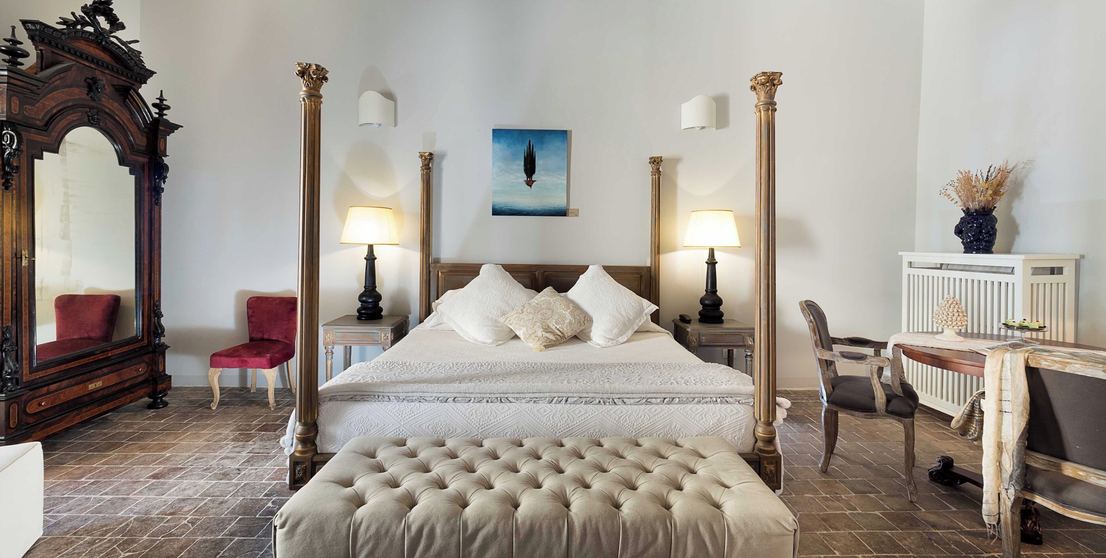 A four-poster bed with a tufted ottoman, a red chair, a wooden desk, and a mirror.