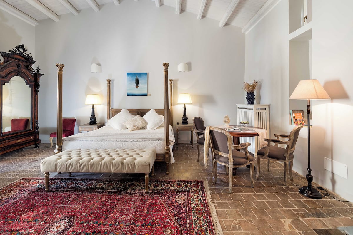 A spacious bedroom with a large bed, a sitting area with a table and chairs, a mirror, and a red patterned rug.