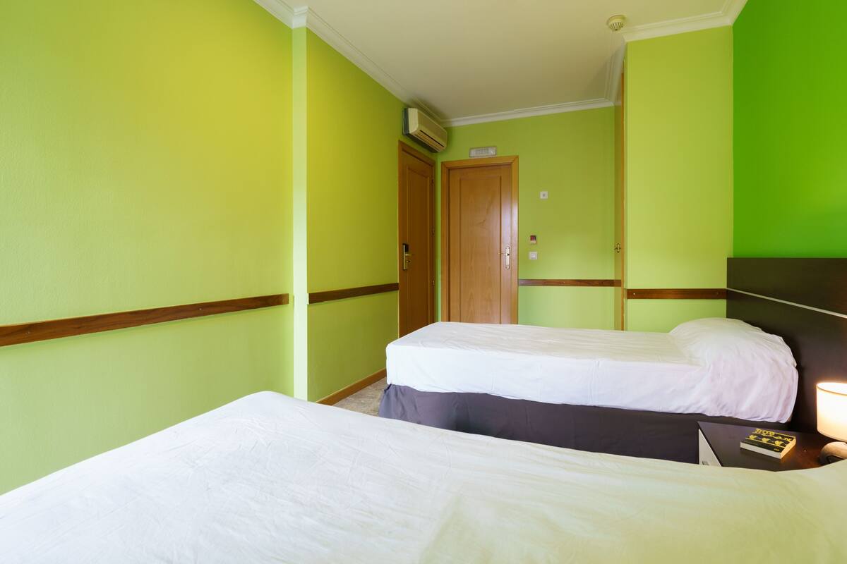 Habitación de hotel con dos camas, cabecera de madera, mesita de noche con lámpara y puerta con un cartel.