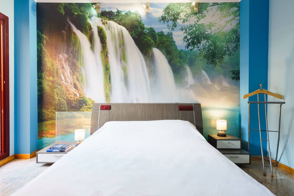 Habitación de hotel con un gran mural de una cascada, una cama con ropa blanca, mesitas de noche con lámparas y un perchero de madera en un soporte.