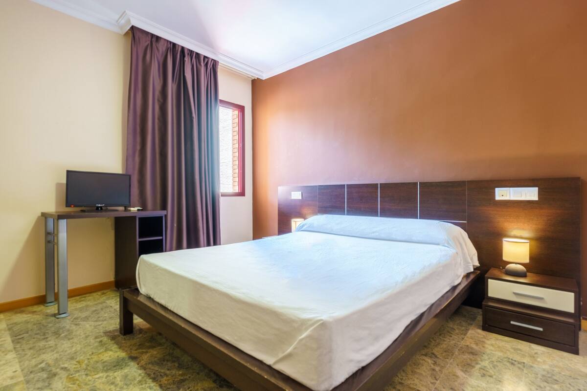 Una habitación de hotel con cama, escritorio con televisor, mesita de noche con lámpara y ventana con cortinas.