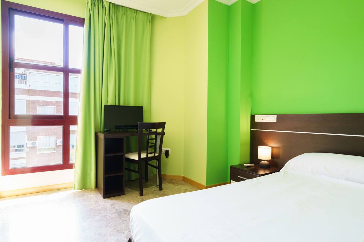 Habitación de hotel con una cama, un escritorio con silla, un televisor y una mesita de noche con lámpara.