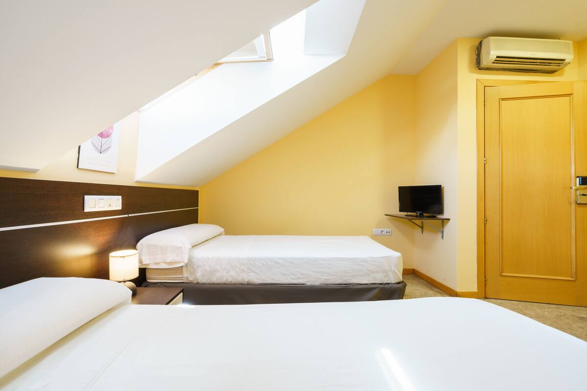 Habitación de hotel con techo inclinado, una cama, una mesita de noche con lámpara, un televisor montado en la pared y una puerta.