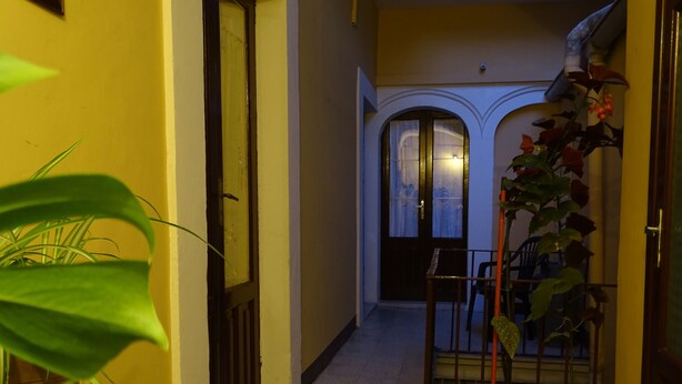 Ein schmaler Flur mit einer hölzernen Bank, Topfpflanzen und einer Tür zu einem Zimmer mit Fenster und Leuchte.