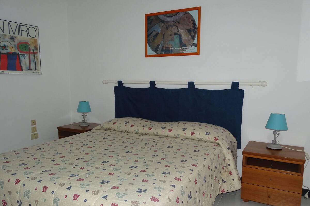 Un dormitorio con cama, dos mesitas de noche, cabecera azul, edredón floral y un cuadro en la pared.
