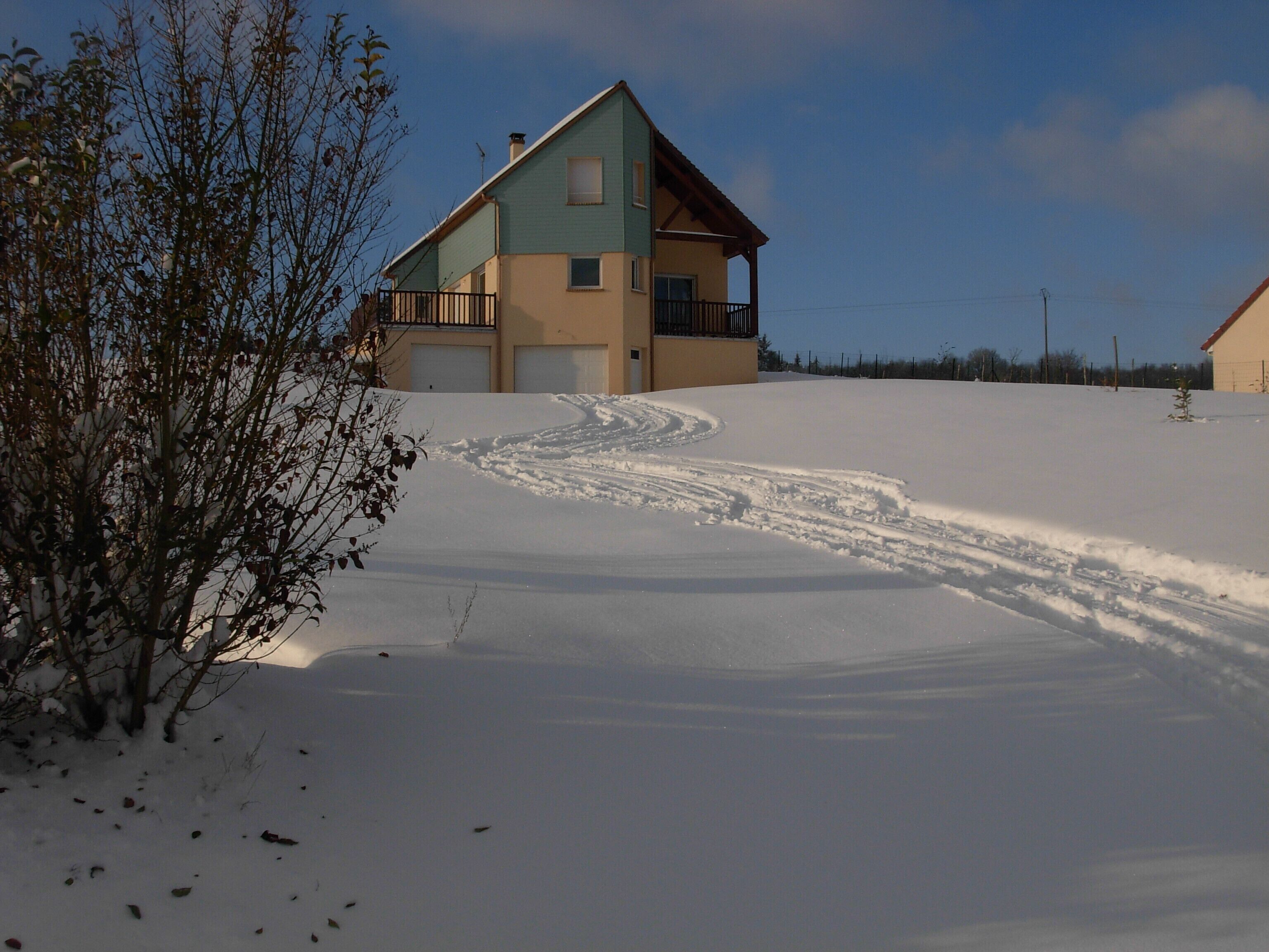 Una casa de dos pisos con un camino de entrada cubierto de nieve y un pequeño árbol en primer plano.