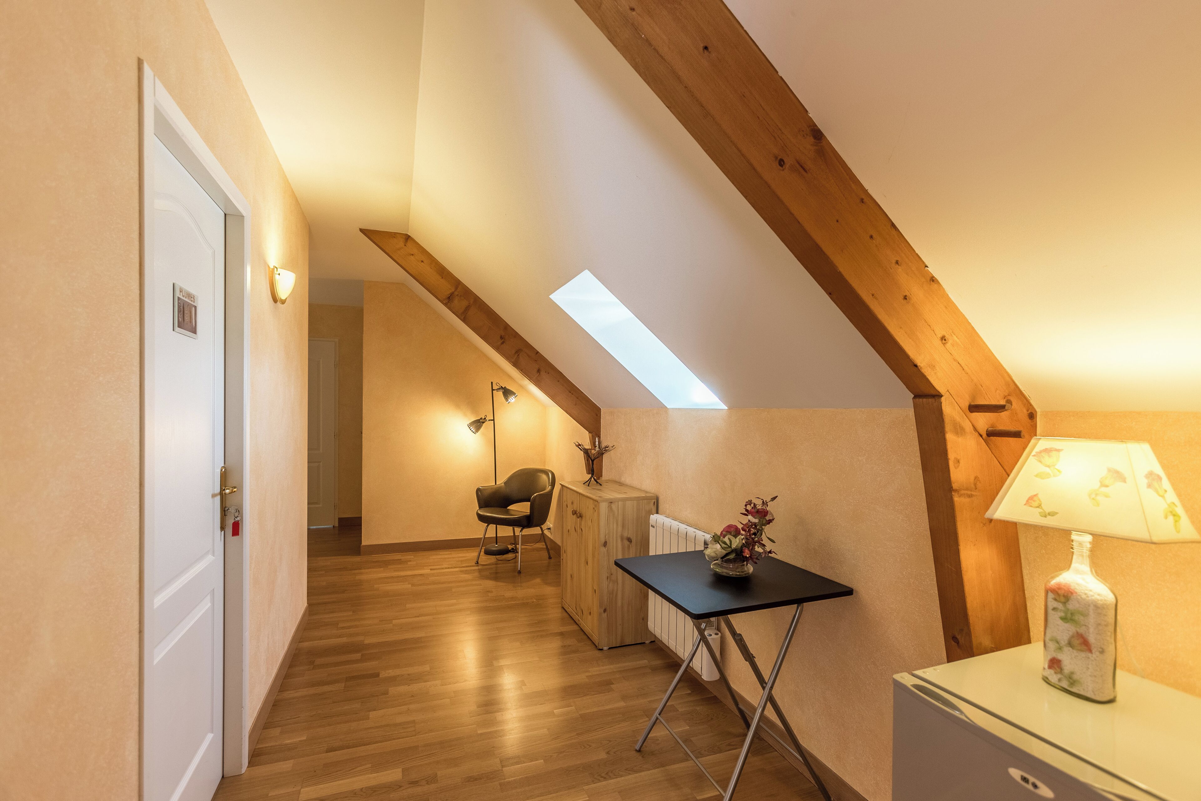 A room with a sloped ceiling, wooden beams, a skylight, a table with a lamp, a chair, and a radiator.