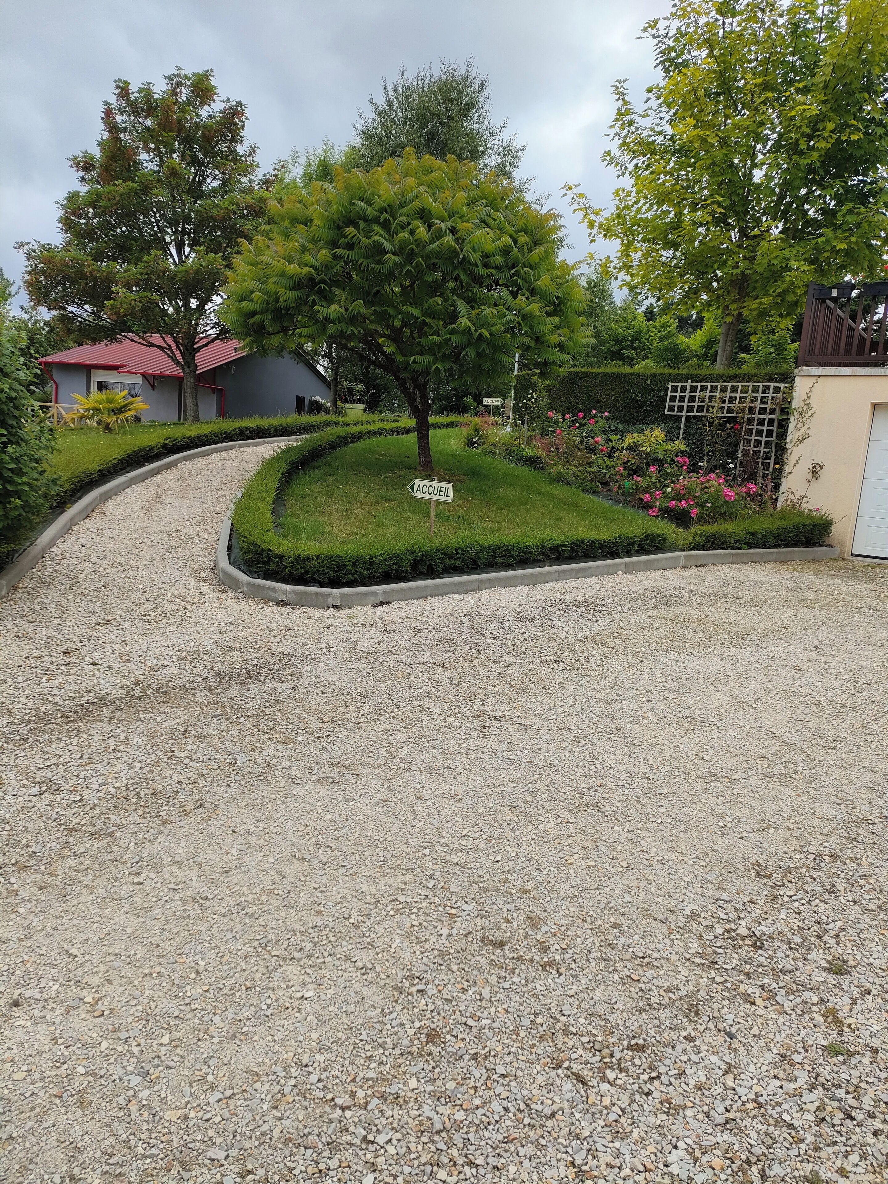 A gravel driveway leading to a house with a red roof, surrounded by well-maintained hedges and flowering plants.