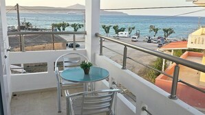 Un balcón con mesa y sillas, con vistas a la costa, coches aparcados y edificios.