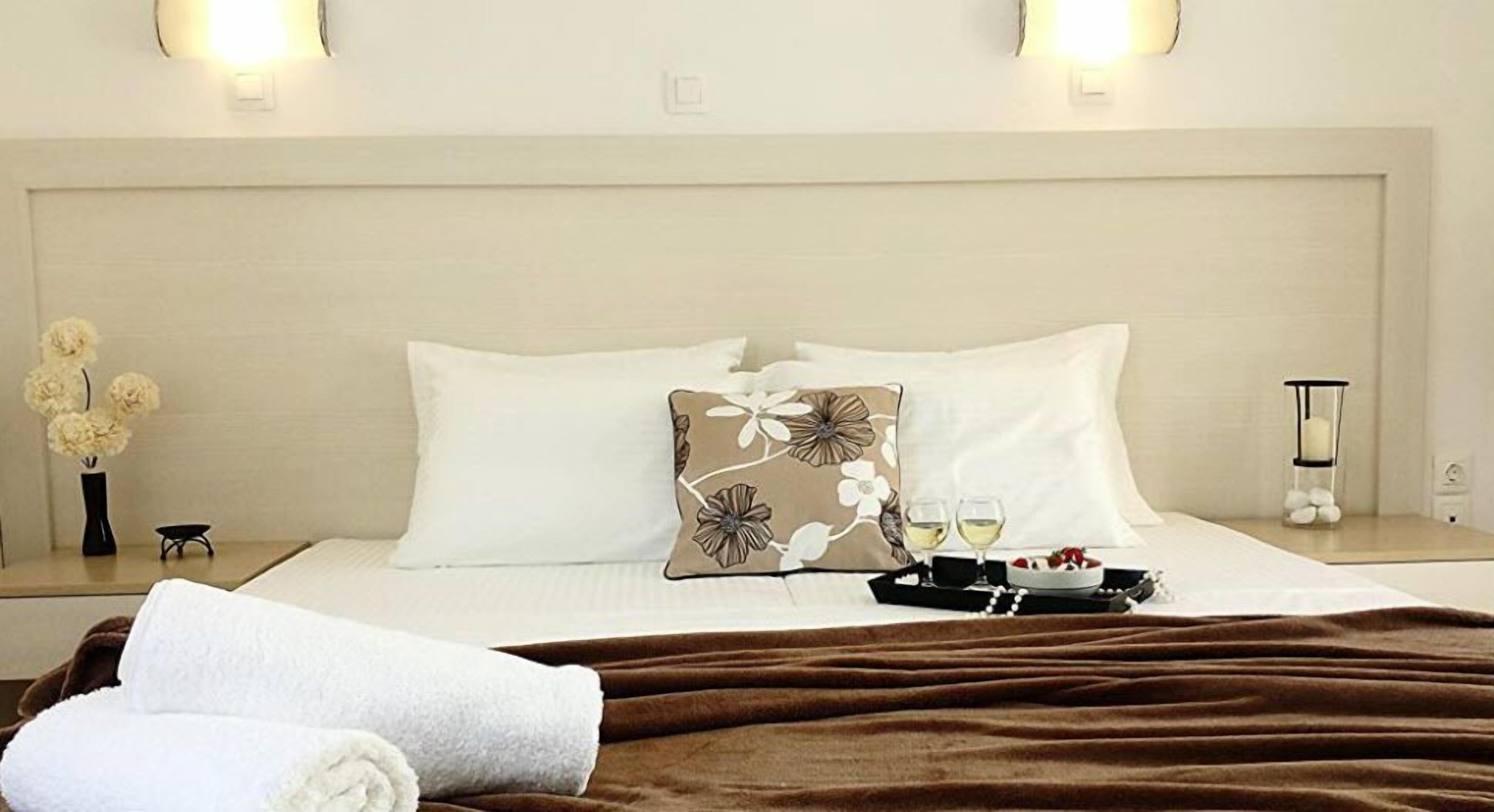 A neatly made bed with a brown blanket, white pillows, and a floral-patterned pillow. There are two glasses of white wine and a small dessert on a tray beside the bed.