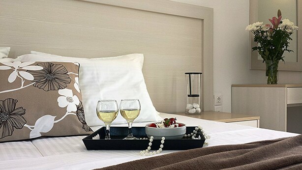 Una cama impecablemente arreglada con una bandeja que contiene dos copas de vino blanco, un cuenco de fresas y una cadena de perlas.