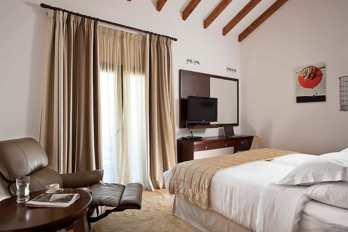 A hotel room with a bed, a desk with a laptop, a TV, a chair, and a window with curtains.