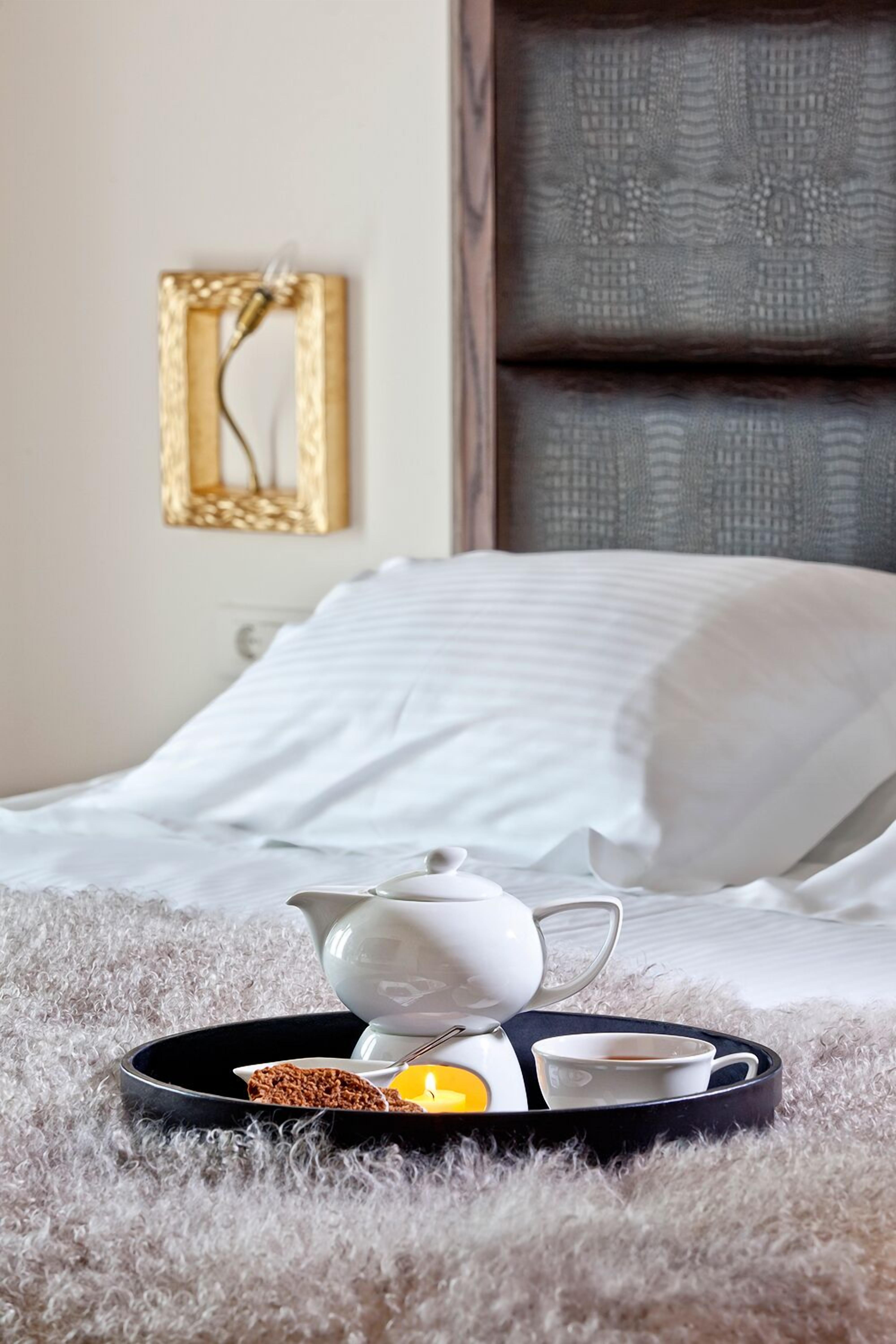 A neatly made bed with white linens, a black tray with a teapot, cups, and a candle on a fluffy rug.
