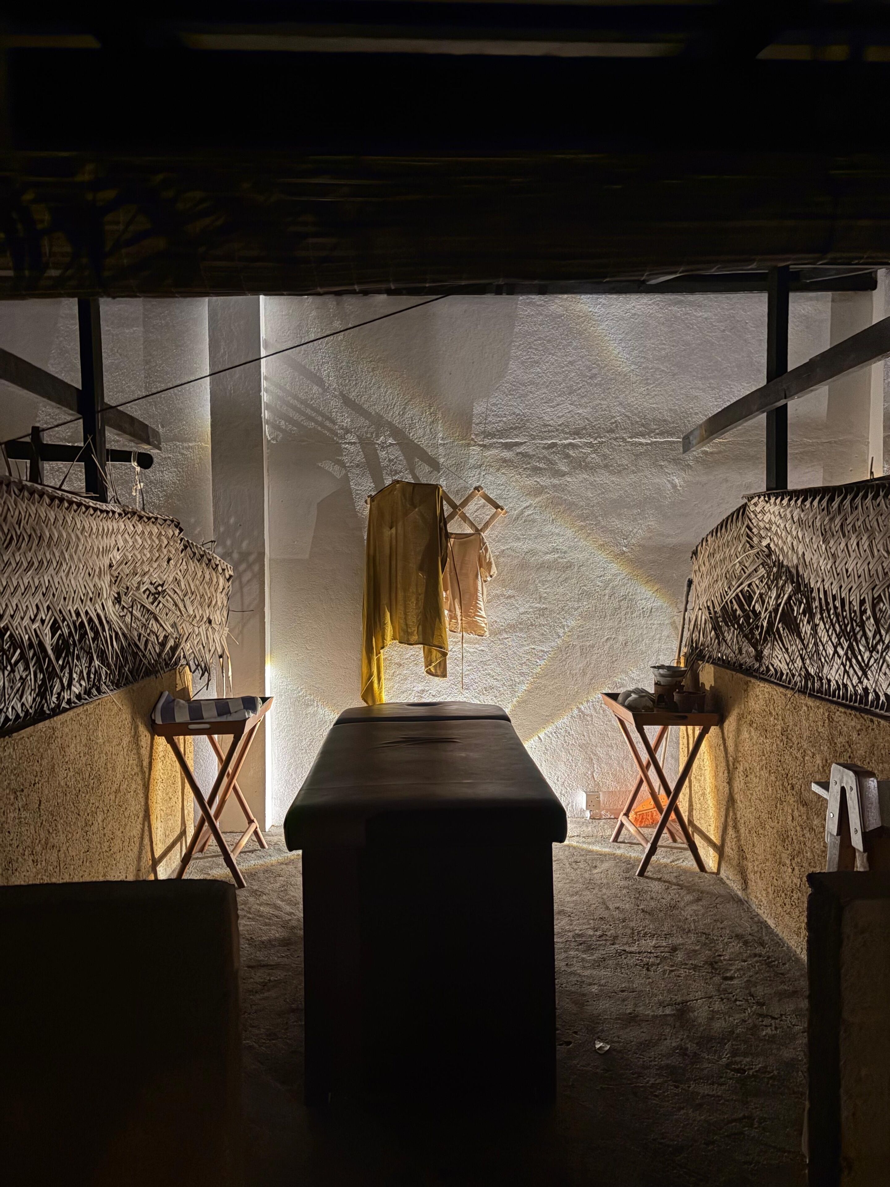 A rustic room with a central bench, two woven wall hangings, and a hanging garment.